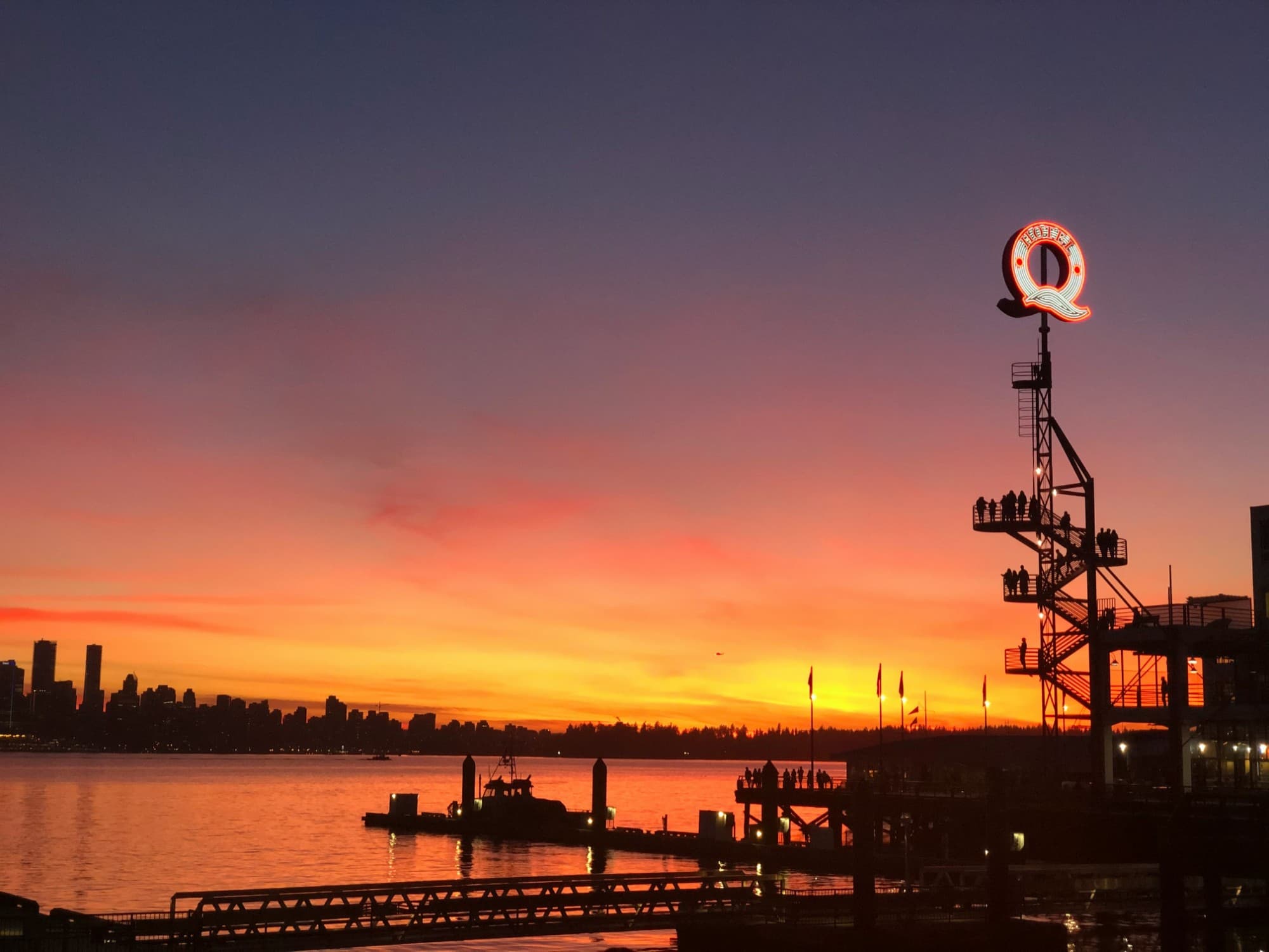 Sunset over the harbour with the Vancouver skyline in silhouette and a lit neon Q sign on a tower